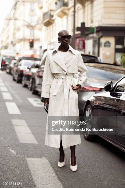 Adot Gak wears silver earrings, a white sweater, a white trench coat with a belt and sunglasses outside the Akris fashion show during the Womenswear...