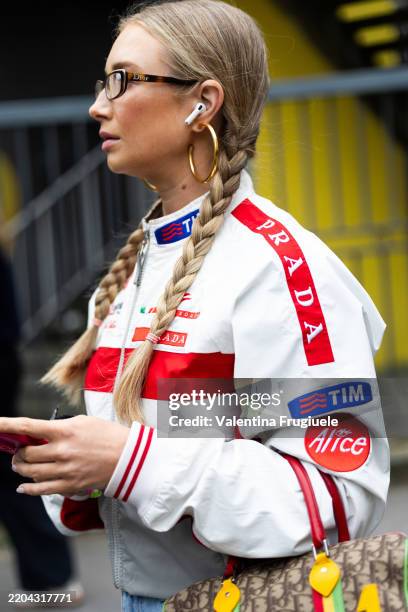 Guest wears Dior glasses, blue jeans, gold hoop earrings, braided hair, a white and red jacket and a Dior vintage bag outside Ottolinger show during...