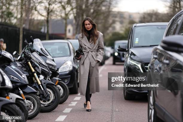 Guest wears black turtleneck long sleeve sweater, light gray belted trench coat, shiny black leather bag, black footless leggings, shiny black...
