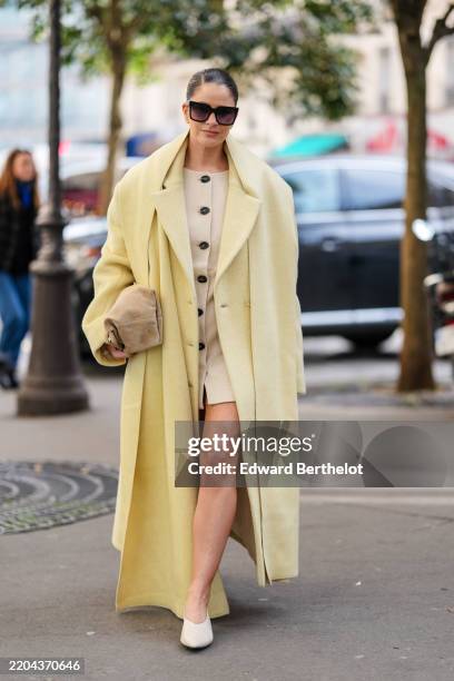 Gili Biegun wears black sunglasses, cream buttoned blazer jacket, light yellow oversized double breasted wool coat, dark yellow scarf, light brown...