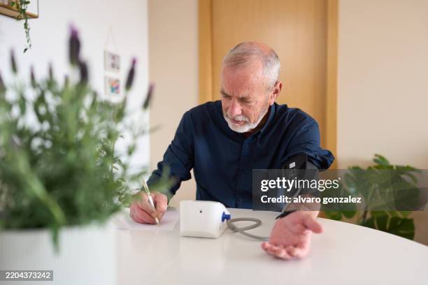 senior man taking blood pressure at home and writing results - hypertensive stock pictures, royalty-free photos & images
