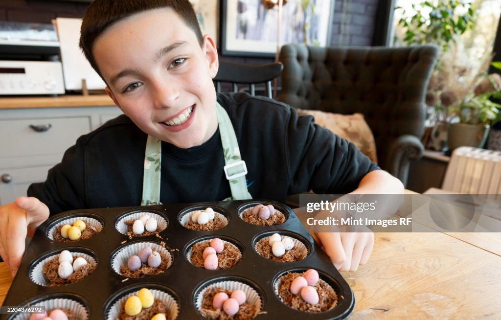 Smiling teenage boy in kitchen presenting freshly made tray of cup cakes to camera