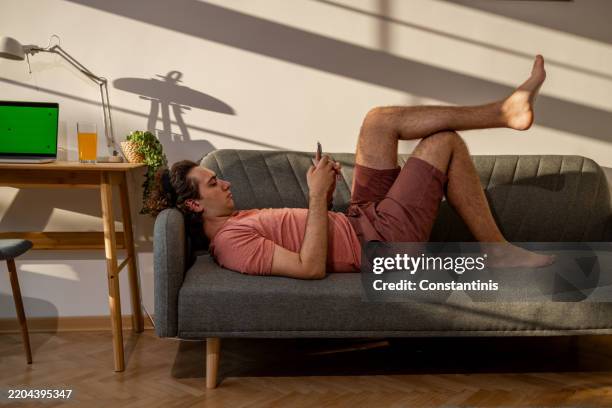 student lying on a sofa in his apartment and using mobile phone - sofa bed stock pictures, royalty-free photos & images