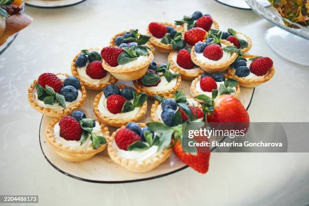 assorted berry tarts with fresh fruits displayed on a white tablecloth - obsttorte stock-fotos und bilder