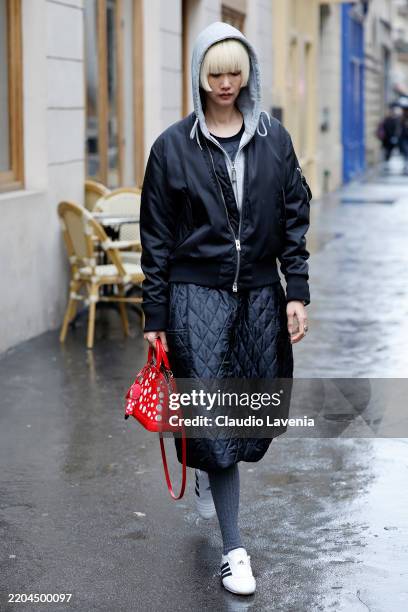 Guest wears grey sweatshirt, black jacket, black quilted skirt, grey socks, white Adidas sneakers, red Louis Vuitton polka dot bag, outside Sacai,...