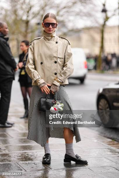 Guest wears dark brown sunglasses, gold earrings, light brown high neck double breasted trench jacket, gold bracelets, gold rings, white black...