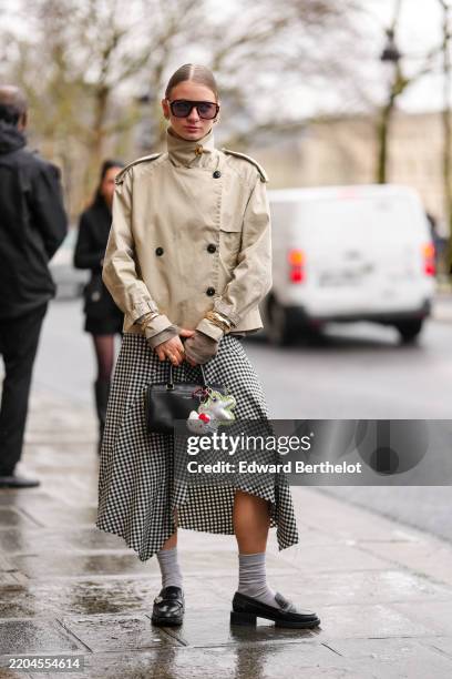 Guest wears dark brown sunglasses, gold earrings, light brown high neck double breasted trench jacket, gold bracelets, gold rings, white black...
