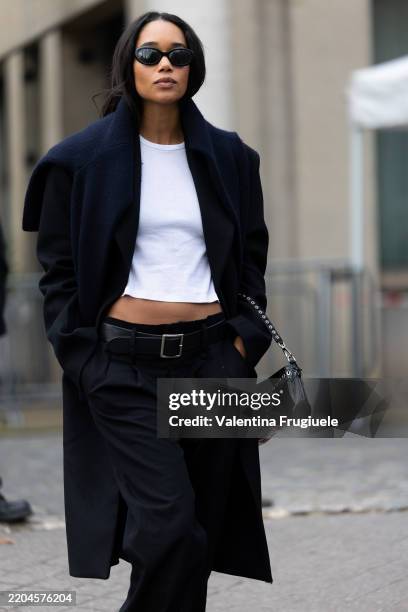 Laura Harrier wears black sunglasses, a blue cardigan, a white cropped top, a black long coat, black wide leg low waist trousers, a black leather...