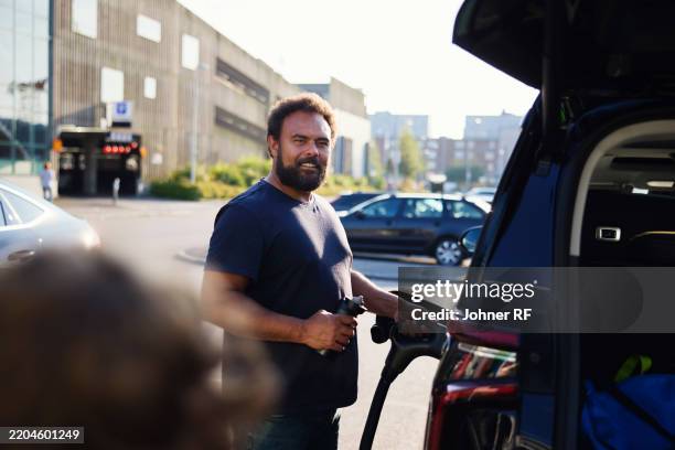 bearded man charging electric car with plug at station on street - hybrid car stock pictures, royalty-free photos & images