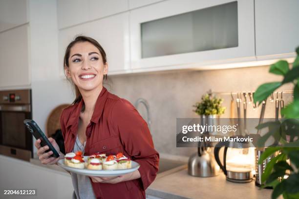 lächelnde frau, die einen teller mit vorspeisen hält und das smartphone in der küche benutzt - mozzarella stock-fotos und bilder