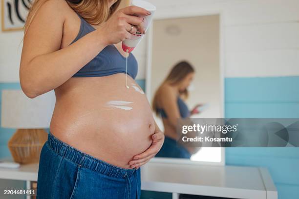 unrecognizable pregnant woman standing in front of mirror holding and applying cream on pregnant belly for moisturizing skin at home - stretch mark stock pictures, royalty-free photos & images