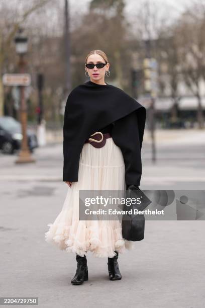 Guest wears black sunglasses, silver hoop earrings, black cape, shiny black silver detail leather belt, white sheer mesh tulle midi skirt, shiny...