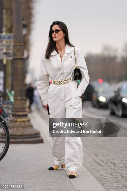 Guest wears black sunglasses, white long sleeve jumpsuit, silver chain belt, shiny black Chanel leather bag, mixed metal rings, light brown black toe...