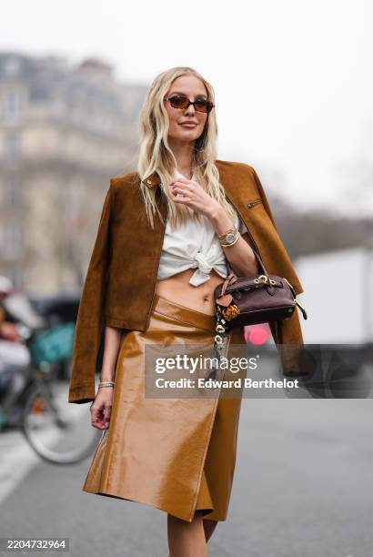 Leonie Hanne wears dark brown sunglasses, white collar tied Miu Miu shirt, dark brown unzipped Miu Miu suede jacket, gold Cartier Bracciale Juste...