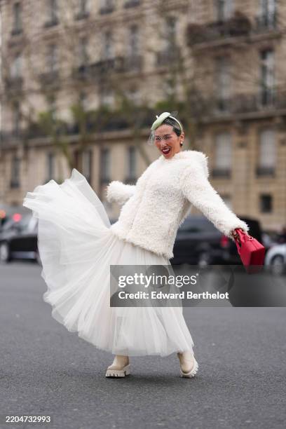 Xuan Xu wears light green hat veil, pearl earrings, cream faux fur zipped oversized hooded coat, white sheer mesh maxi skirt, shiny burgundy bordeaux...