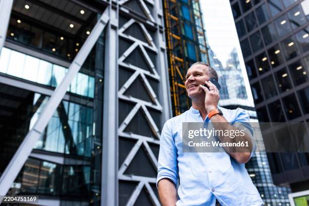 smiling businessman talking on smart phone in front of buildings - angle photos et images de collection