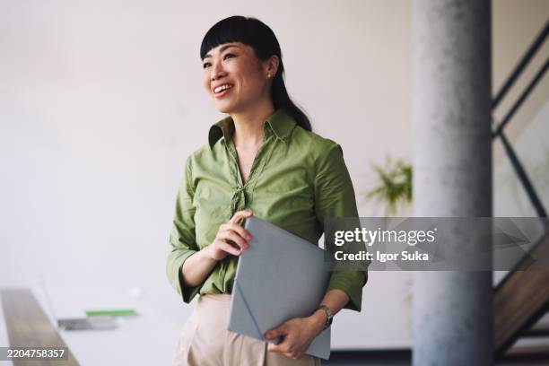 smiling japanese businesswoman holding a laptop and looking away in a modern office - looking away stock pictures, royalty-free photos & images