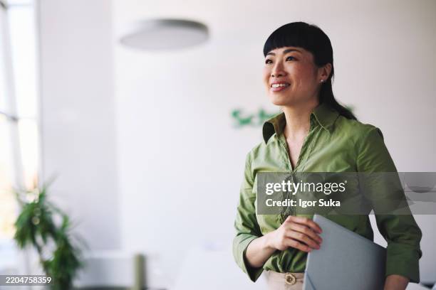 japanese businesswoman holding laptop and looking away smiling in office - simple green stock pictures, royalty-free photos & images