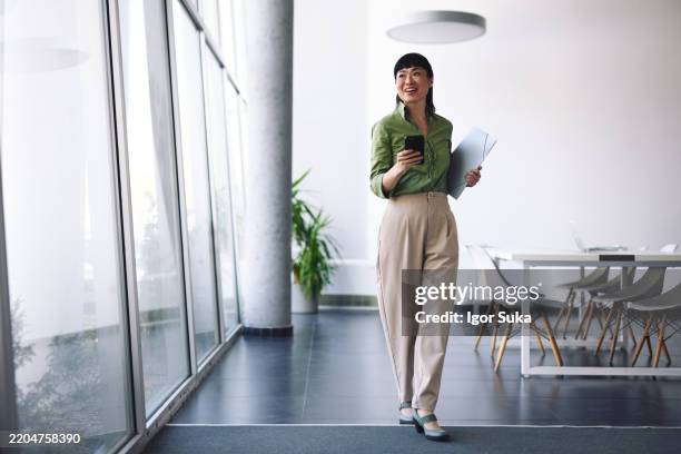 giovane imprenditrice asiatica che cammina in un ufficio moderno con documenti e smartphone - figura intera foto e immagini stock
