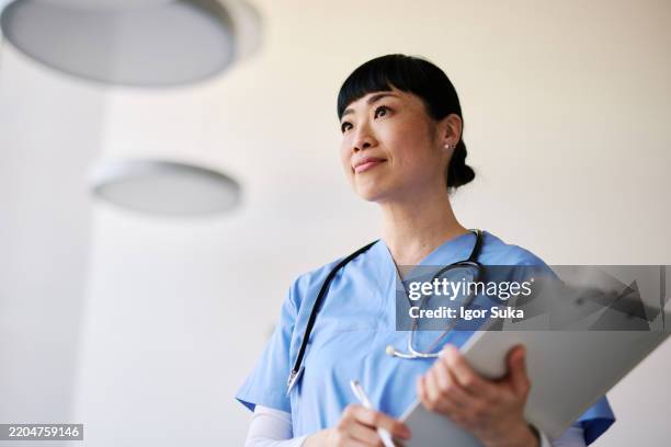 japanese nurse looking up holding a clipboard and pen in a hospital - female likeness stock pictures, royalty-free photos & images