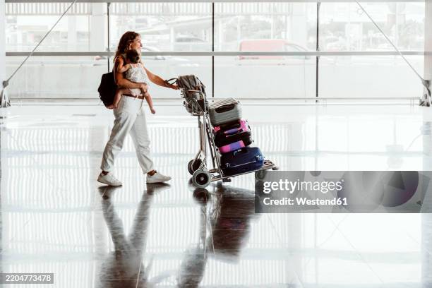 happy woman carrying baby daughter and pushing luggage cart at airport - luggage trolley stock pictures, royalty-free photos & images