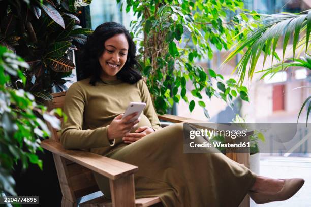 pregnant woman using mobile phone and sitting on chair in balcony - maternity dresses stock pictures, royalty-free photos & images