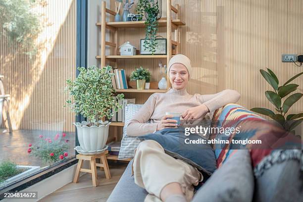 young adult female cancer patient wearing headscarf sitting on sofa , relaxing. - cancer radiation stock pictures, royalty-free photos & images