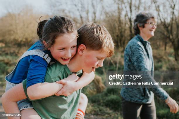 a brother carries his little sister on his back as their grandmother supervises them in the background - middle childhood stock pictures, royalty-free photos & images