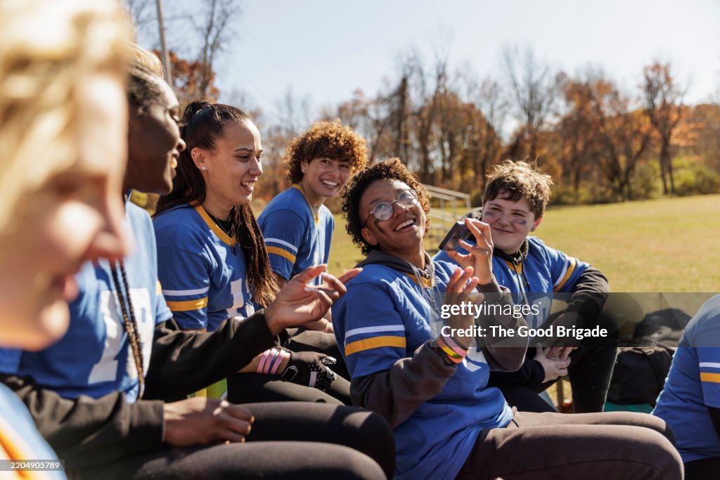 Happy curly haired football player using smart phone with friends while sitting bleachers