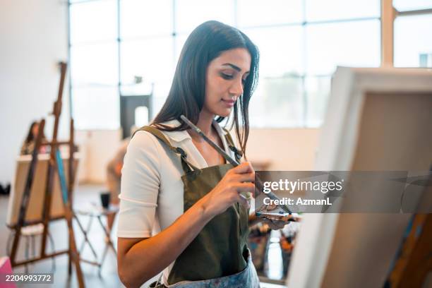 focused middle eastern female artist painting in bright studio with natural light - easel stock pictures, royalty-free photos & images