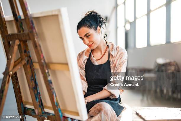joven artista femenina del medio oriente que pinta en un estudio iluminado por el sol brillante - lienzo de artista fotografías e imágenes de stock