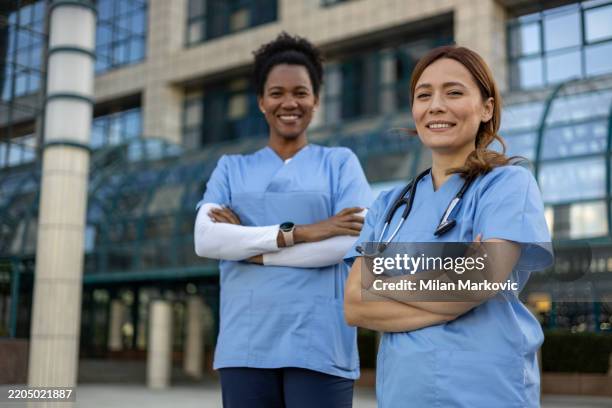 two smiling female doctors standing outside hospital with arms crossed - serviços-essenciais imagens e fotografias de stock