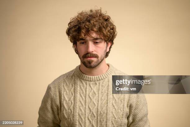 junger mann mit lockigem haar, der zopfstrickpullover vor beigem hintergrund trägt - schüchtern stock-fotos und bilder
