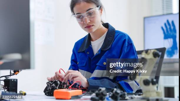 ingegnere donna bianca che indossa occhiali protettivi di sicurezza con giacca da lavoro blu sta saldando intricati componenti robotici su un circuito stampato o un'officina di dispositivi elettronici in una stanza di laboratorio - ordine foto e immagini stock