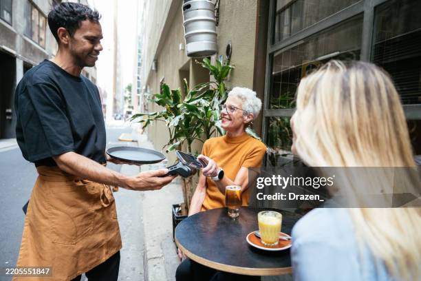 bartender serving two women in cozy cafe. woman paying with contactless credit card on pos device - aboriginal and torres strait islander commission stock pictures, royalty-free photos & images