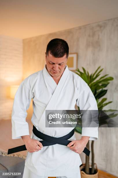 mid-adult man fastening his black blet while wearing kimono and practicing karate at home - fastening stock pictures, royalty-free photos & images