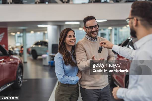 happy couple receiving new car keys from salesman in dealership - autokredit stock-fotos und bilder