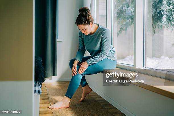 woman suffering from knee pain while sitting on windowsill at home - gonfio foto e immagini stock
