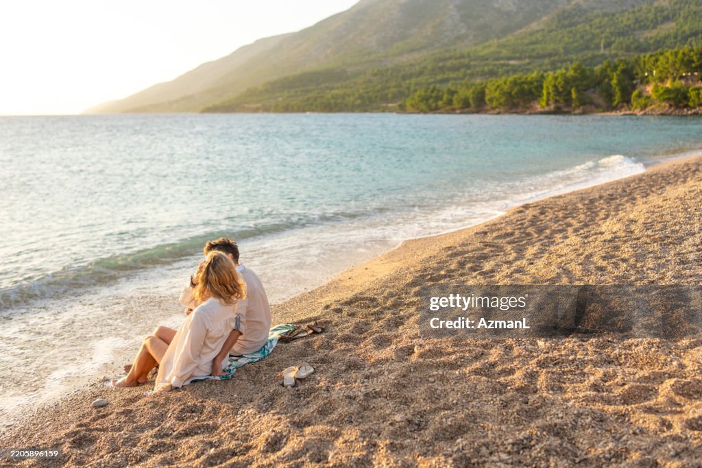 Mid-Aged Couple in White Clothing Sitting Together on Sandy Beach at Golden Hour
