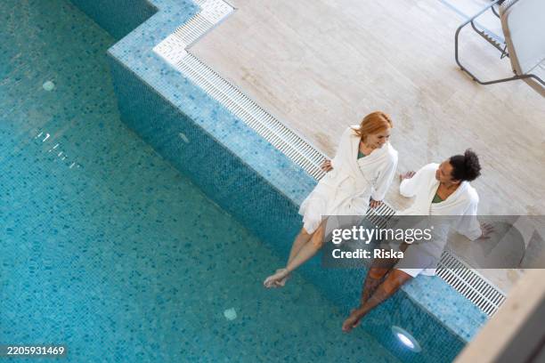 two women in white robes relaxing by the spa pool - spa weekend stock pictures, royalty-free photos & images
