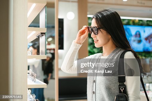 Young Asian woman shopping for glasses in optical store looking at mirror. Making a choice for buying a new pair. Young Asian woman shopping for glasses in optical store looking at mirror. Making a choice for buying a new pair.