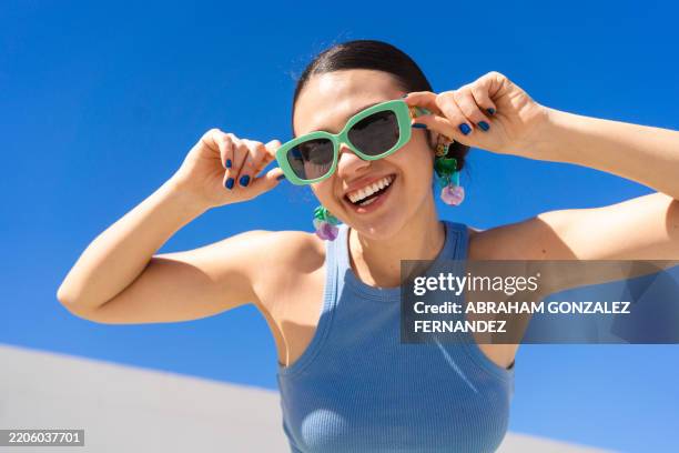 cheerful woman in sunglasses beaming brightly under cloudless azure sky - brillen en lenzen stockfoto's en -beelden