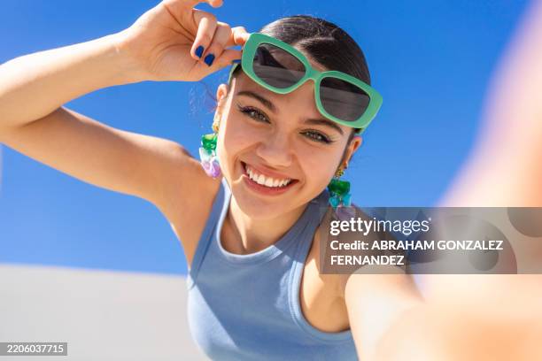 smiling woman capturing selfie, adjusting stylish sunglasses with bright blue sky backdrop - solglasögon bildbanksfoton och bilder