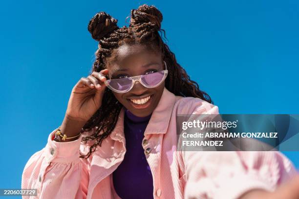 fröhliche frau mit geflochtenem haar, die die sonnenbrille senkt und ein strahlendes lächeln unter einem wolkenlosen azurblauen hintergrund enthüllt - sonnenbrille stock-fotos und bilder