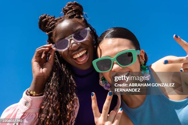 tres jóvenes alegres con gafas de sol sonríen y hacen un gesto con el signo de la paz contra un cielo azul claro - gafas de sol fotografías e imágenes de stock
