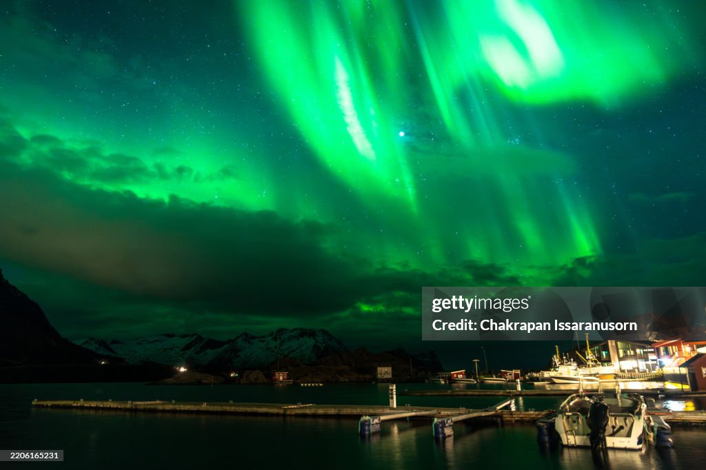 Massive Northern lights (aurora borealis) KP3 appearing green over the sky at a dock with boats in Senja island, Norway