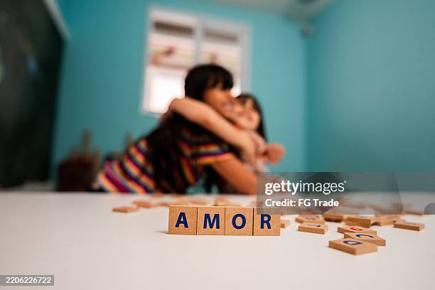 primer plano de un bloque de juguete con alfabeto en portugués y español (amor) y hermanas abrazándose en el fondo - una palabra fotografías e imágenes de stock