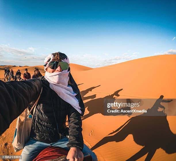young man takes selfie on camel safari in sand dunes - dromedaris stockfoto's en -beelden