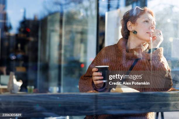 woman contemplating at cafe window. 48-year-old caucasian with coffee cup gazes thoughtfully outside. - me time stock pictures, royalty-free photos & images