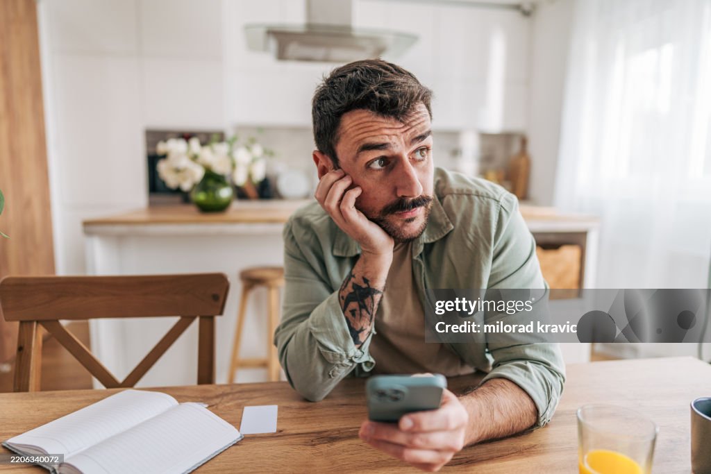 Pensive man holding smartphone and looking away while sitting at table